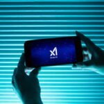 A person holds a telephone displaying the logo of Elon Musk s artificial intelligence grok in front of a background lit by a blue light.