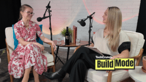 Two women sit facing each other in a podcast studio, speaking into microphones. One wears a red floral dress and glasses, while the other wears a cream top and black pants. A small table between them holds a bottle of wine, glasses of water, and books. A TechCrunch Build Mode logo appears in the corner.