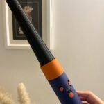 Mão segurando o MultiStyler e Secador Dyson Airwrap id, um modelador de cachos com corpo preto, alça azul e laranja e pequenos botões na alça