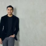 A well-dressed man leans against a neutral colored wall